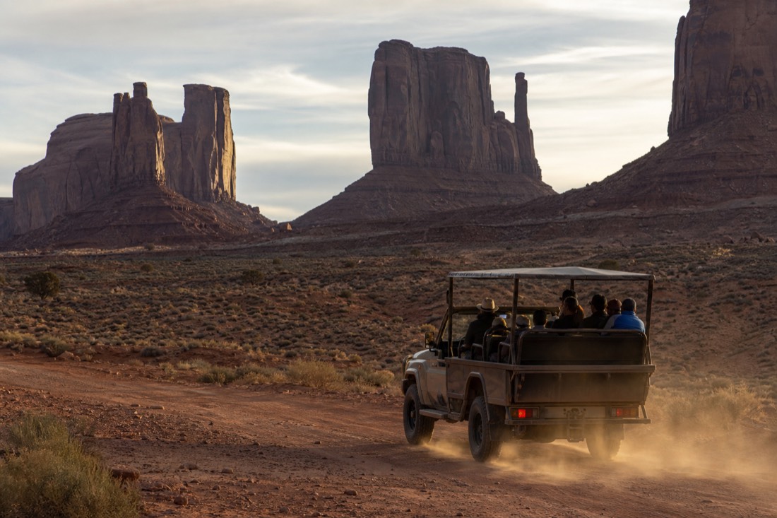 An open-air 4x4 with a Navajo guide and small group driving the red dirt valley floor below the sandstone buttes of Monument Valley